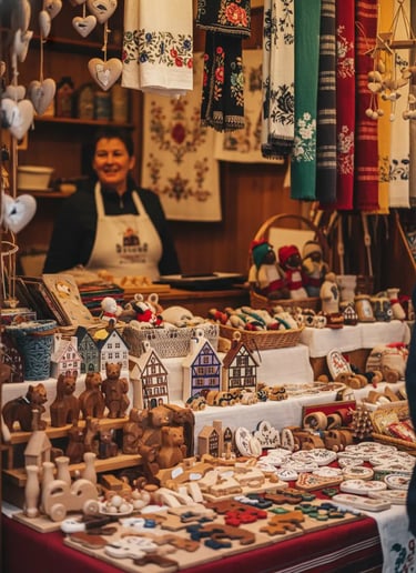 A smiling vendor at a Christmas market stall piled high with various handmade goods and small souven