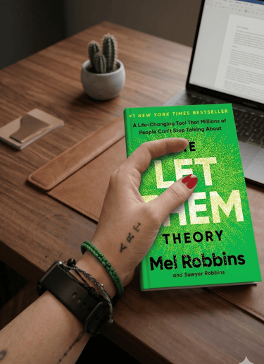 A person holding The Let Them Theory book by Mel Robbins at a wooden desk with a laptop.