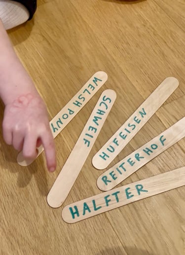 Child pointing at wooden craft sticks with horse-related German words on a floor.