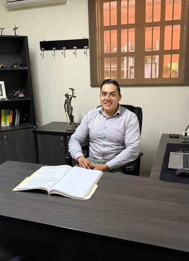 A smiling professional lawyer sitting at a dark wood desk in an organized office with law books and a laptop.
