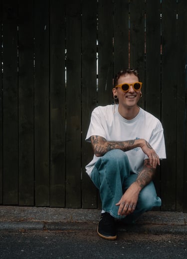 Cody with yellow sunglasses, a baggy t shirt, and baggy jeans, crouched in front of a green fence.
