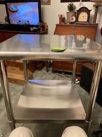 A compact stainless steel table with an adjustable undershelf positioned in a home living room near a television.