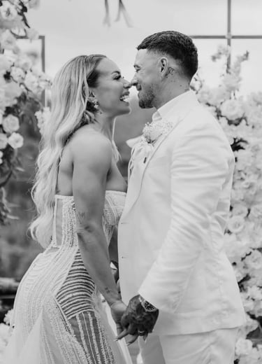 Smiling bride and groom in white wedding attire holding hands at an outdoor floral ceremony.