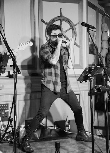 Singer Leo Couto performing on stage with a microphone, wearing a hat, black jeans, a plaid shirt, and sunglasses.