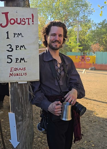 Man in pirate costume holding a tankard and camera next to a Joust! sign at a fair