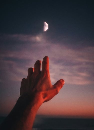 Hand reaching toward a crescent moon in a dark evening sky with sunset clouds.