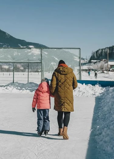 A parent and a young child in a pink jacket holding hands while walking onto a sunny outdoor ice ska