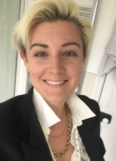 smiling blonde woman, blue eyes, dark eyebrows, wearing a white shirt, black jacket and gold chain