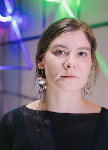 Das Portrait einer jungen Frau, die gerade in die Kamera schaut. Im Hintergrund bunte Lampen.
