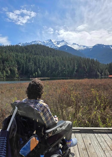 Sean in his power wheelchair on a lakeside boardwalk looking at Mt Currie in BC