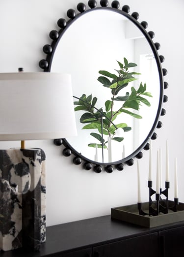 Modern black bead round mirror reflecting a fiddle leaf fig plant next to a marble table lamp.