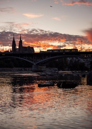 Sunset over Basel city skyline with a tram crossing the bridge near Basel Minster cathedral.