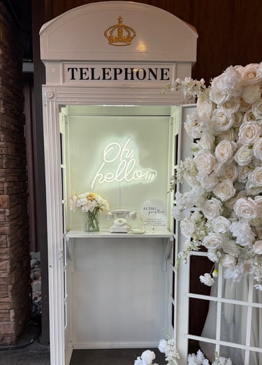 Vintage white telephone booth with floral arrangements and neon sign for wedding guestbook audio recording.