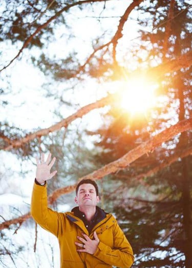 Person in yellow jacket with hand raised, standing in sunlit snowy forest with tall pines.