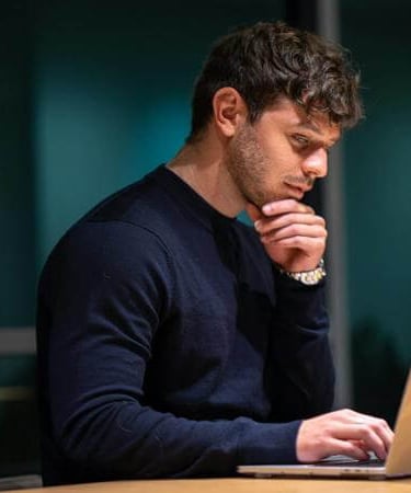 Individual seated at table with hand on chin, concentrating on a laptop in a dimly lit room.