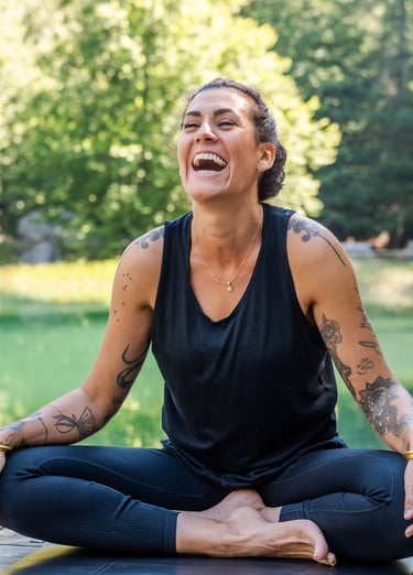 A tattooed woman laughing while practicing outdoor yoga in a cross-legged seated pose.