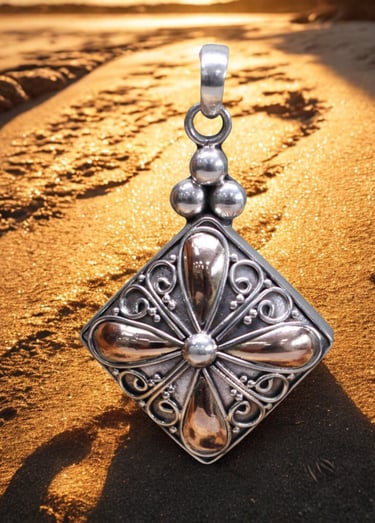 Handcrafted silver and bronze filigree pendant resting on golden beach sand during sunset.