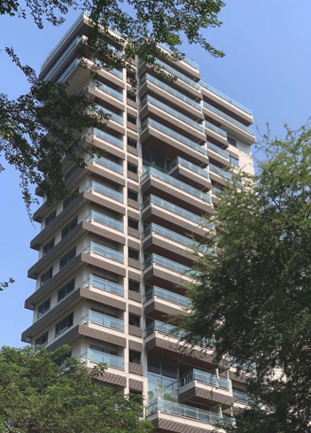 Modern luxury high-rise apartment building with glass balconies viewed through green trees.