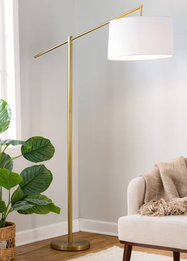 A modern gold arched floor lamp with a white drum shade standing next to a cream armchair.