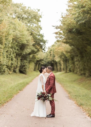 A bride in a white lace dress and a groom in a red suit kissing on a tree-lined road.