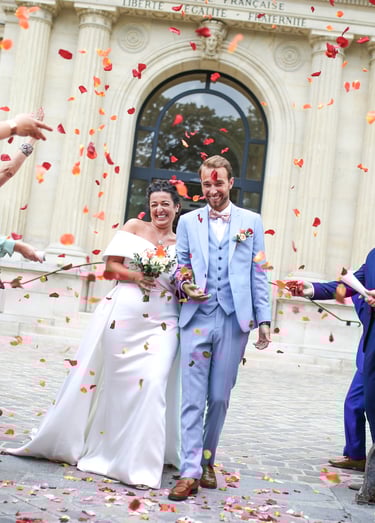 Smiling bride and groom exit a French building while guests toss orange rose petal confetti.