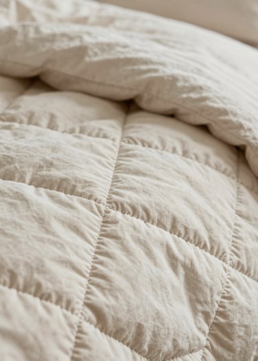 A close-up photograph showing the intricate hand-stitched patterns on a soft cream and warm taupe quilt. The lighting is soft and natural, coming from the side to highlight the raised texture of the fabric. The composition is a detailed macro shot of the stitching.