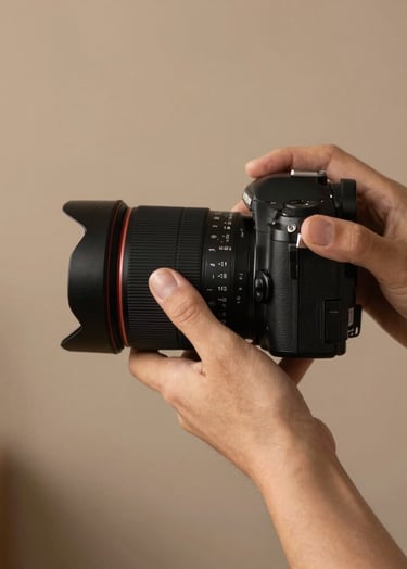 A close-up photograph of a professional photographer's hands adjusting a camera lens, set against a minimalist North American studio backdrop. The lighting is soft and warm, highlighting the textures of the equipment and the charcoal black and earthy brown tones of the surrounding space.