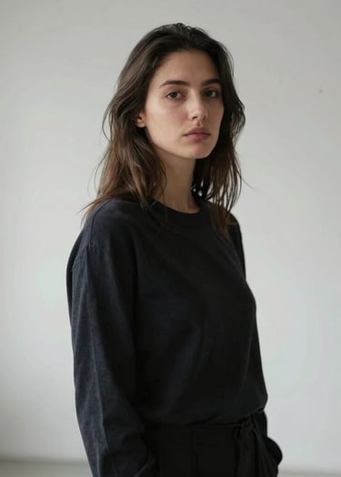 A sophisticated artistic portrait of a woman in a minimalist French studio. She wears simple, dark attire and looks thoughtfully past the camera. The background is a clean white smoke wall with soft, natural lighting creating a serene mood.