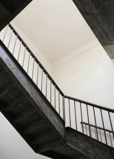 A low-angle perspective shot looking up through the geometric structure of the steel stairs. The dark charcoal lines intersect against a soft off-white ceiling, highlighting the mathematical beauty of the fabrication.