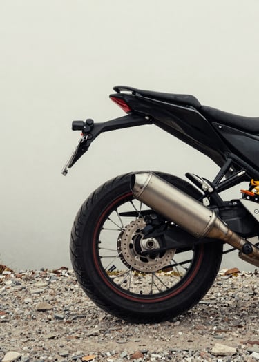 A minimalist composition of a motorcycle's rear wheel and exhaust pipe resting on a gravel path in a Global / Western highland. The background is a soft off-white foggy sky.