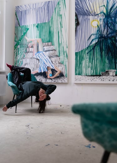 Artist posing upside down in a teal chair in front of large abstract contemporary paintings in a studio.