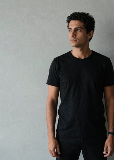 A portrait of a creative professional in a South American / Brazilian setting, dressed in minimalist black attire, standing against a light gray textured wall with soft side lighting.