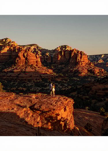 A wide, cinematic landscape photograph of a couple standing on a ridge in Sedona, Arizona, during sunset. The North American / US environment is flooded with warm orange and charcoal shadows, capturing a grand yet intimate moment.