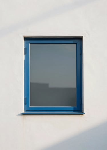 A minimalist architectural photograph of a single steel blue window frame against a soft cloud white wall. The composition is clean and centered, emphasizing the geometric purity and the soft play of morning shadows.
