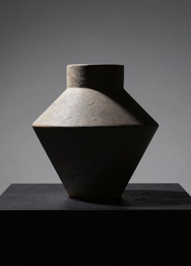 A minimalist still life of a single geometric ceramic sculpture on a matte black pedestal. The lighting is focused and dramatic, creating sharp silhouettes against a soft gray backdrop. Sophisticated North American / European gallery aesthetic.