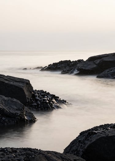 A minimalist, artistic landscape of the Brest shoreline in Brittany. The composition is clean, featuring dark charcoal rocks meeting a misty off-white sea. The lighting is soft and ethereal, reflecting a serene and sophisticated mood.