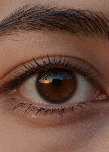A poetic close-up of a person's eye, capturing a sharp reflection of a sunset over the sea. The image is emotional and soulful, with rich natural tones of warm sand and deep charcoal in the iris and lashes.