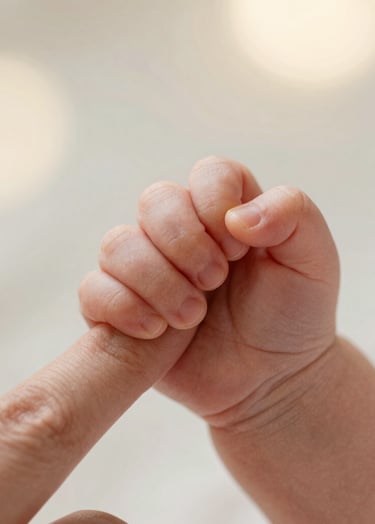 A detailed close-up shot of a newborn baby's tiny hand holding a parent's finger, photographed outdoors with very soft, creamy bokeh and warm morning light. Simple, emotional, and visually clean.
