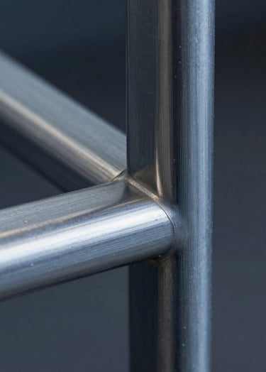 Macro detail of a brushed aluminum joint on a designer chair. The metal has a dusty grey-blue sheen, and the craftsmanship is evident in the seamless connection. The background is a soft-focus deep charcoal.