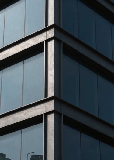 A minimalist photography shot focusing on the corner of a modern building. Muted slate steel beams meet dusty grey-blue glass panels. The lighting is sharp, creating dramatic deep charcoal shadows that emphasize the building's skeletal frame.