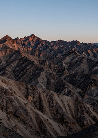 Wide-angle landscape of a rugged mountain range in Northeast Nevada during the blue hour. The peaks are sharp and clear against a tranquil, pale sky. The composition conveys profound tranquility and vastness, using sand and dark brown tones.