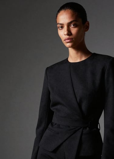 An editorial fashion portrait of a Brazilian model wearing a structured black outfit. Minimalist studio setting with dramatic side lighting and deep shadows. Professional, clean, and artistic style.