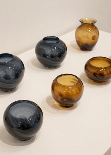 Minimalist photography of a series of artistic glass forms on a Soft Off-White surface in a Northern European / Welsh / British gallery. Each form contains swirls of Deep Charcoal and Warm Ochre, captured with high-resolution clarity and clean composition to evoke curiosity and innovation.