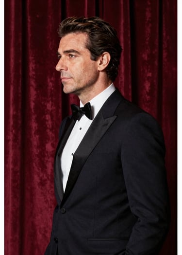 A sophisticated profile portrait of a world-famous Italian tenor in a midnight black tuxedo, standing before heavy Borgogna red velvet stage curtains. Soft side lighting highlights his features, creating a deeply artistic and elegant atmosphere with a palette of #262626 and #6B242D.