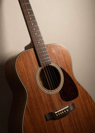A close-up photograph of a professional acoustic guitar made of dark cocoa brown wood, leaning against a soft cream-colored studio wall. The lighting is warm and cinematic, highlighting the grain of the wood and the silver strings.