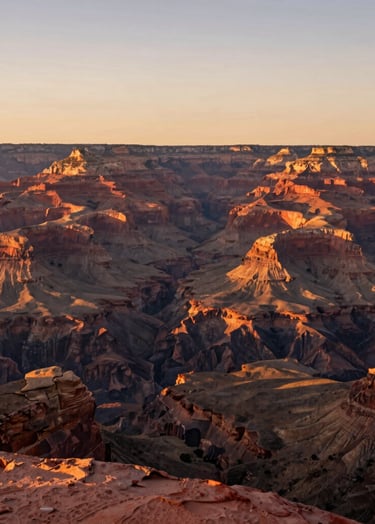 A wide cinematic shot of a majestic North American / US canyon at golden hour. The landscape is bathed in warm light, highlighting the vibrant terracotta rock formations against a soft sand sky. The composition is clean and breathtaking, focusing on the scale of the natural environment.