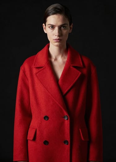 Minimalist high-fashion portrait of a subject wearing a bold red structured coat, dark black background, dramatic rim lighting from the side, North American studio setting, sleek professional photography.