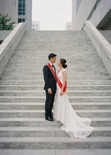 An editorial-style photograph of a couple standing on a minimalist concrete staircase in a North American / US downtown area, the bride wearing a red bridal sash, overcast soft lighting.