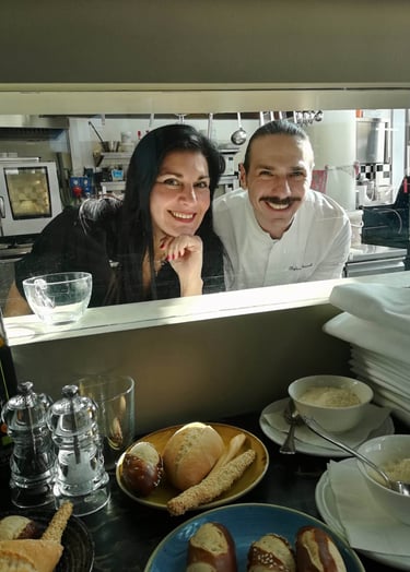 Clara Mennella e uno chef affacciati al pass della cucina, tra pane fresco e mise en place, simbolo del racconto culinario.