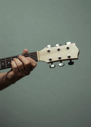 A high-contrast photograph of a musician's hand adjusting a tuning peg on a guitar headstock. The style is raw and authentic, with a soft off-white light hitting the hand and a muted slate green wall in the background.